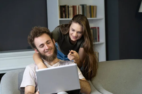 Δουλεύοντας μαζί νεαρό ζευγάρι που μένει σε καραντίνα στο σπίτι με να κάθεται στις πολυθρόνες. Ευτυχισμένο νεαρό ζευγάρι κάθεται σε μια καρέκλα μαζί χρησιμοποιώντας το laptop. Τετράγωνα — Φωτογραφία Αρχείου