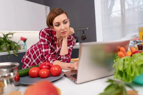 Femme aux cheveux courts utilisent ordinateur portable ayant appel vidéo près des légumes frais dans la cuisine. Femme ennuyée ou intéressée utilise un ordinateur portable. Jeune femme à la cuisine à la recherche en ligne pour des recettes d'aliments sains — Photo