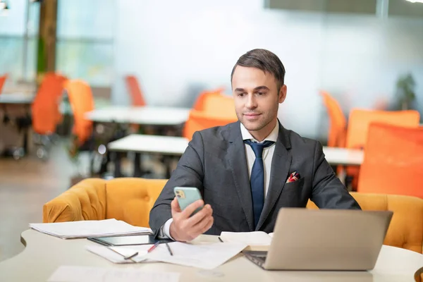 Sonriente joven empresario o gerente mirando por teléfono mientras trabaja en el ordenador portátil sentado en la oficina moderna con sillas naranjas tóxicas en el fondo — Foto de Stock