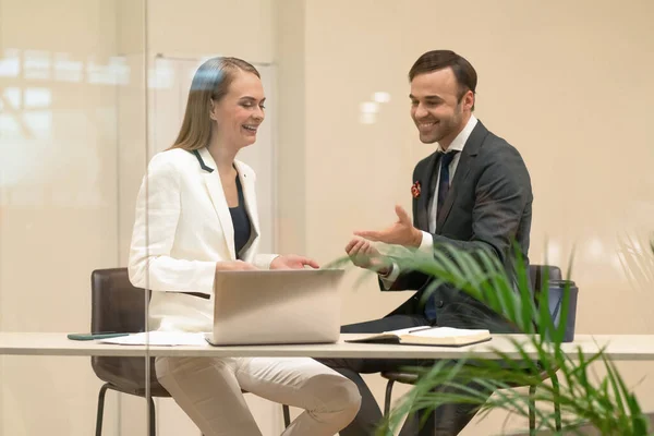 Två skrattande projektledare diskuterar uppdateringar samtidigt som de tar en fikapaus. Unga affärsmän i kostym med kaffe i händerna står bakom glaset — Stockfoto