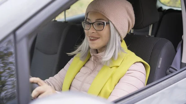 В машині їде азіатка. Зріла жінка в окулярах, дивлячись на дорогу з рульовим колесом. Блондинка середньолітня азіатка в машині. Жінка на дорозі. Концепція бізнесу — стокове фото