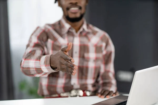 Afroameričan na volné noze natahuje ruku pro koktejl sedí v kanceláři s notebookem. Mladý podnikatel potřásá rukou návštěvníkovi, který sedí v kanceláři. Koncept pohovoru — Stock fotografie