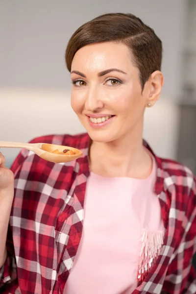 Close up dona de casa em camisa xadrez vermelho e sopa de cabelo curto cozinhar na cozinha moderna. Mulher sorridente a testar o seu prato. Chef mulher se divertindo na cozinha — Fotografia de Stock