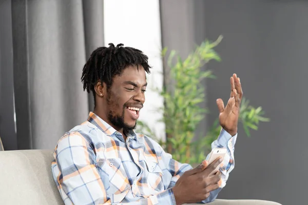 Glad skrattande afroamerikansk student använda smartphone med videosamtal eller titta på online-videor. Porträtt av glad man gestikulera titta på smartphone sitter på soffan. Begreppet sociala medier — Stockfoto