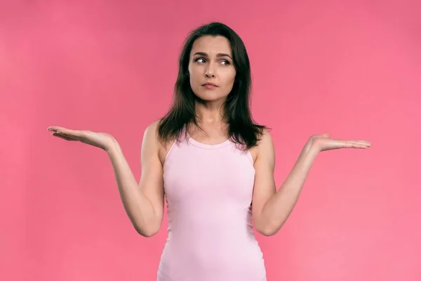 Balancing or weighting up young woman in pink female sleeveless T-shirt and jeans looking at camera with thoughtful look isolated on pink background — Stock Photo, Image