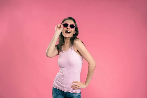 Sexy happy girl in sun glasses kiss shaped wearing tank top and denim jeans posing in studio isolated on pink background — Stock Photo, Image