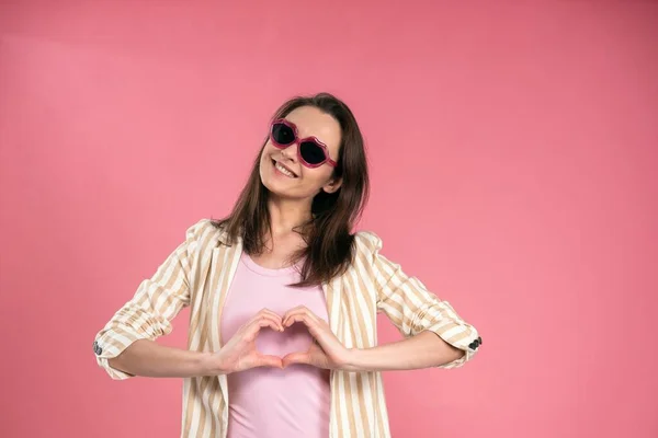 Щаслива бізнес-леді в сонцезахисних окулярах поцілунок у формі показуючи знак любові з руками у формі серця, одягнені в смужку куртку позує в студії ізольовані на рожевому фоні — стокове фото