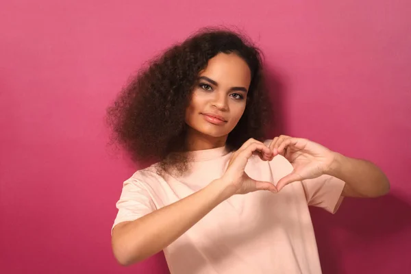 Εμφάνιση σημάδι αγάπης Αφροαμερικανός κορίτσι με μακριά μαλλιά μπούκλα κοιτάζοντας κάμερα φορώντας peachy t-shirt απομονώνονται σε σκούρο ροζ φόντο. Έννοια αγάπης — Φωτογραφία Αρχείου