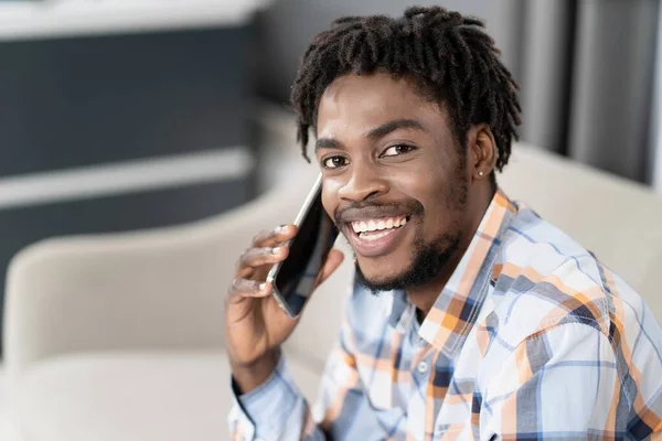 Ung afroamerikansk kille med telefon i handen tittar på kameran medan du pratar. Porträtt av lycklig man som har närvarande samtal sitter på soffan. Begreppet sociala medier — Stockfoto