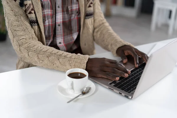 Uomo afroamericano che digita sul computer portatile con una tazza di caffè accanto a lui indossando maglione beige seduto al tavolo della caffetteria del campus. Gocce di caffè su cucchiaio isolato su tavolo bianco — Foto Stock