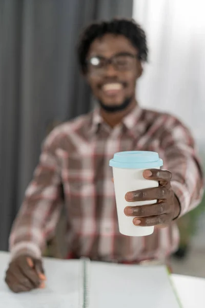 Focus selettivo sulla mano dell'uomo afroamericano con tazza di cartone o tazza usa e getta per caffè o tè. Tazza monouso bianca con piombo di plastica blu nella mano dell'uomo. Concetto piatti usa e getta — Foto Stock
