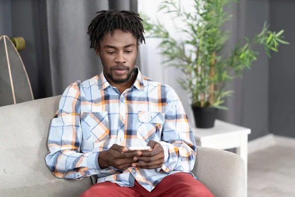 Koncentrerad eller intresserad afroamerikansk man använder smartphone kontrollera sociala medier eller uppdatera information. Porträtt av en ung man som håller smartphone sittande i soffan. Begreppet sociala medier — Stockfoto