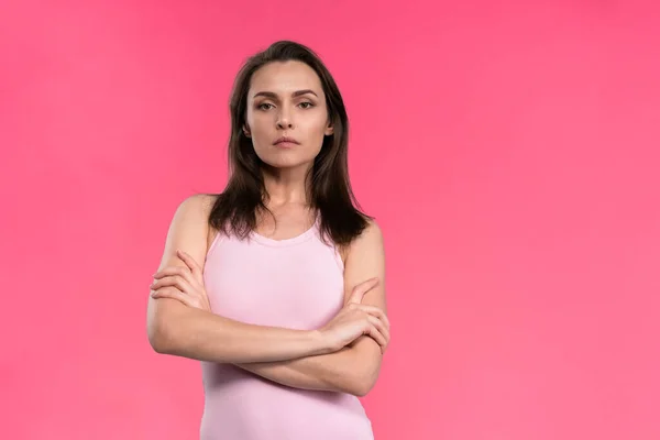 Misstänksam ung kvinna i en rosa ärmlös t-shirt vikta armarna och tittar på kameran. Tvivel, misstro ung vacker kvinna isolerad på rosa bakgrund — Stockfoto