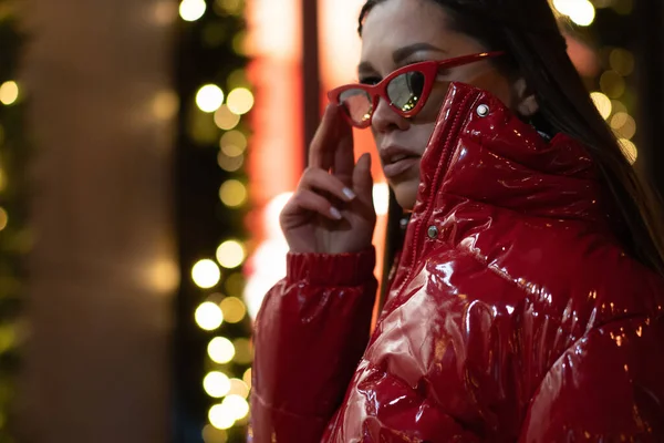 Model gadis mode dalam kacamata matahari dengan rambut dikepang dan mantel mengkilap merah berdiri di luar ruangan di latar belakang kota malam. Konsep kehidupan malam. Konsep kehidupan klub malam — Stok Foto