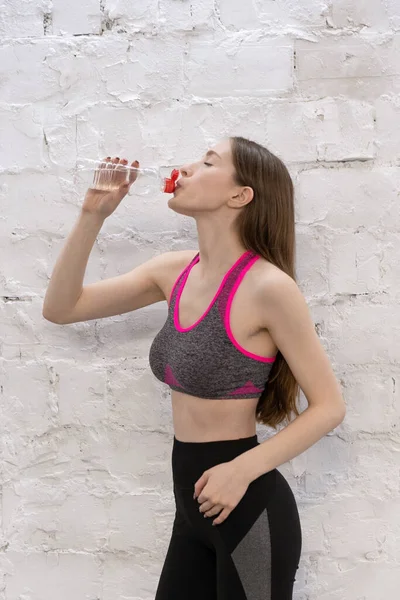 Menina atlética bonita vestindo sportswear bebendo água fazendo bico após o exercício de pé ao lado da parede de tijolo branco. Mulher após desidratação desportiva. Conceito de desporto — Fotografia de Stock