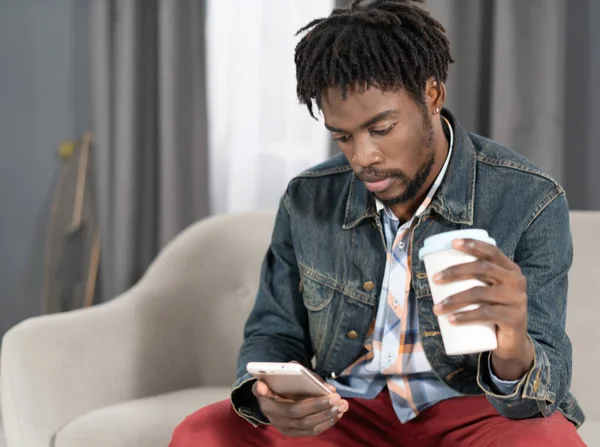African American young man holding paper or disposable cup with plastic lid and smartphone in hand. Portrait of happy man having present conversation sitting on the sofa. Social media concept — Foto Stock