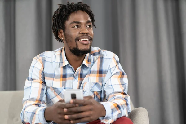 African American young man holding phone or smartphone in hand looking away. Portrait of happy man having present conversation sitting on the sofa. Social media concept — Fotografia de Stock