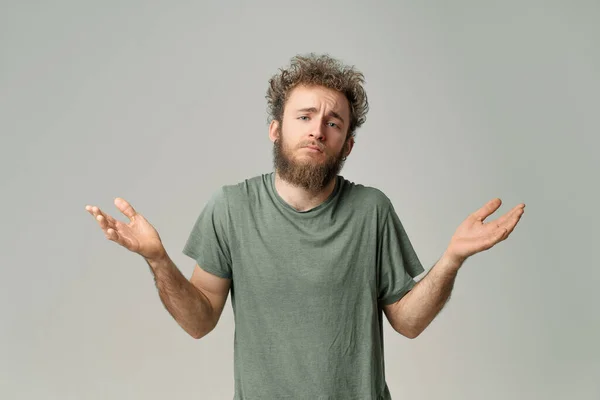 Gesturing I DONT KNOW or I AM SORRY young handsome bearded wild curly hair man with bright blue eyes isolated on grey background. Young thinking man in green t shirt on white. Copy space — Stockfoto