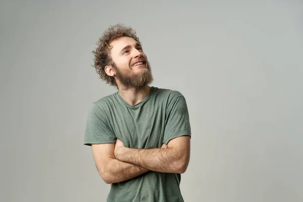 Smiling handsome young man with bearded and wild curly hair, bright blue eyes looking up with hands folded isolated on white background. Young thinking man in green t-shirt on white background — Stockfoto