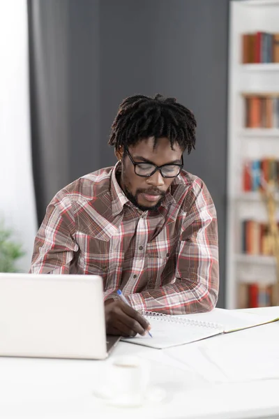 Young african student man making notes working at home working on laptop or listening teacher or boss during video call. African student study using laptop at home — Stockfoto