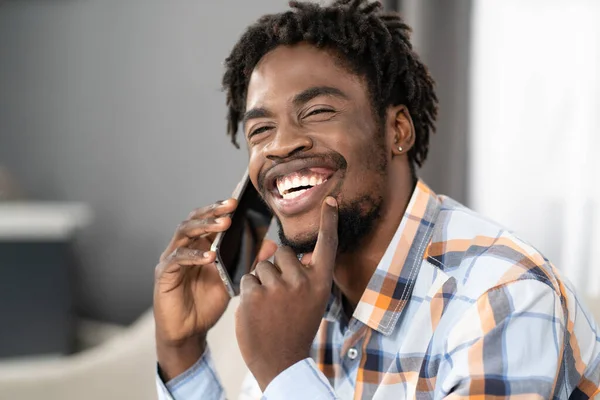 Happy smiling young African American man talking on the phone holding smartphone in hand looking away. Portrait of happy man having present conversation sitting on the sofa. Social media concept —  Fotos de Stock