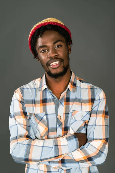 Fiatal, mosolygós fekete férfi afro kalapban, karját összehajtva a kamerába néz. Vidám afro-amerikai fiatalember kockás ingben nézi kamera elszigetelt szürke háttér — Stock Fotó