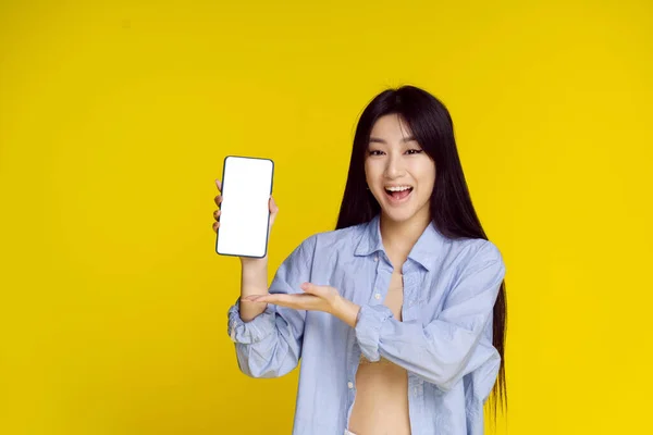 Молода щаслива азіатська дівчина тримає смартфон, показуючи білий порожній екран і захоплююче вигравати ізольовані на жовтому тлі. Чудова пропозиція. Розміщення продукту. Реклама мобільних додатків — стокове фото