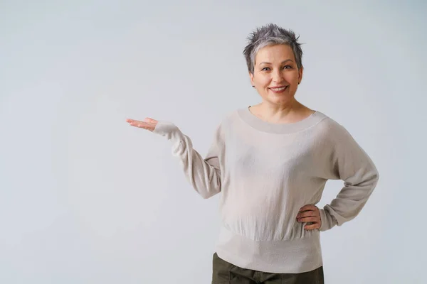 Beautiful, gorgeous 50s mature woman with grey hair posing with hand up for your product placement isolated on white background. Copy space and place for product placement. Aged beauty — стоковое фото
