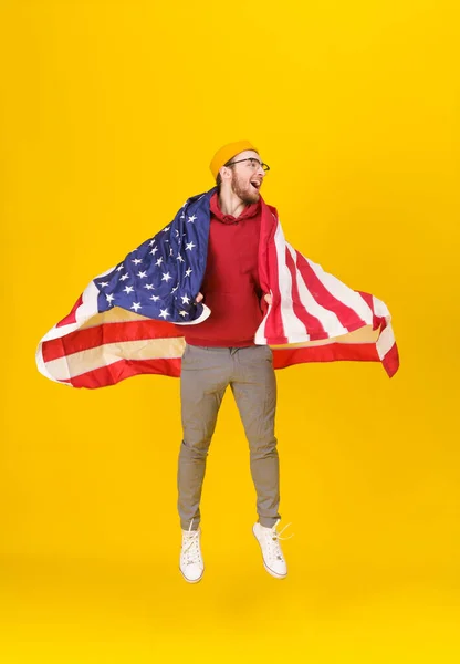 Joven feliz, divertido con capucha roja y bandera de EE.UU. saltando altura aislado sobre fondo amarillo. la libertad está en tu vida. Joven alegre con bandera americana —  Fotos de Stock