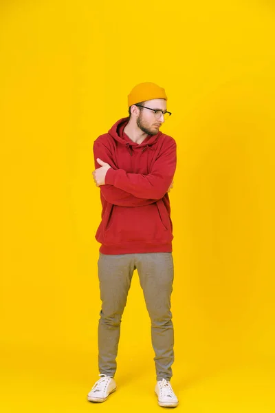 Ein kräftiger junger Mann in Freizeitkleidung mit Brille posiert seitwärts auf gelbem Hintergrund, die Arme verschränkt. Stilvoller bärtiger smarter Hipster-Mann im Casual-Look. Nachdenklicher Typ auf gelbem Hintergrund — Stockfoto