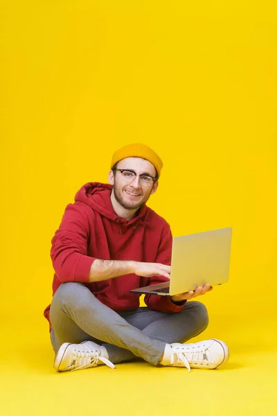 Happy young hipster man in red hoodie sitting on the floor holding laptop studying or working. Smart freelancer caucasian man with laptop isolated on yellow background — Foto de Stock