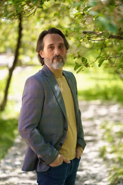 Mature handsome man with grey beard looking thoughtful at the camera wearing casual jacket and jeans holding hands in pockets — Stock Photo, Image