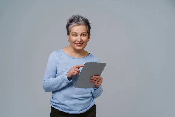Mujer madura de pelo gris sonriente de 50 años sosteniendo tableta digital trabajando o comprobando en las redes sociales. Mujer bonita de 50 años en blusa azul aislada en blanco. Personas mayores y tecnologías. Imagen tonificada — Foto de Stock
