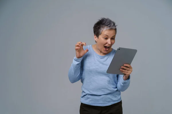 Inspired, shocked mature grey hair businesswoman with digital tablet in hand working, shopping, gaming online. Pretty woman in 50s in blue blouse isolated on white.