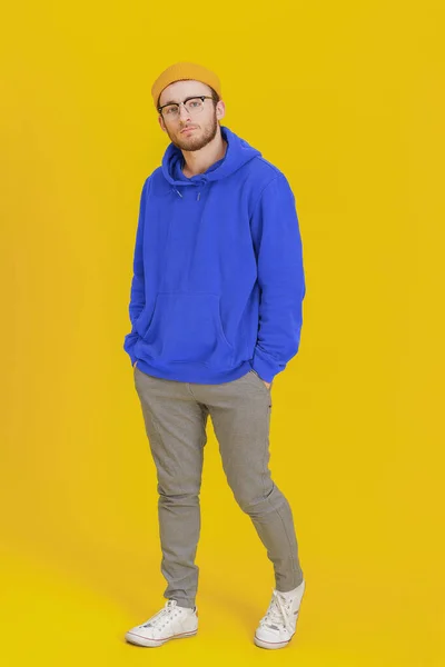 Young man model in casual wear, glasses posing looking at camera on yellow background with hands in pockets. Stylish bearded smart hipster man casual look. Thoughtful guy on yellow background.