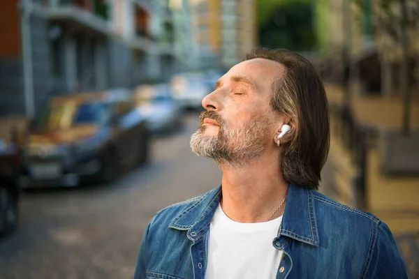 目を閉じて人生を聴いて音楽を楽しむ屋外の古い町の通りハンサムな成熟した灰色のひげを生やした男カジュアル服を着て無線イヤフォン 旅行ライフスタイルのコンセプト — ストック写真