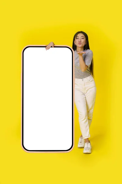 Sending air kisses asian girl stand near huge, giant smartphone with white screen dressed in casual isolated on yellow background. Free space mock up mobile app advertising.