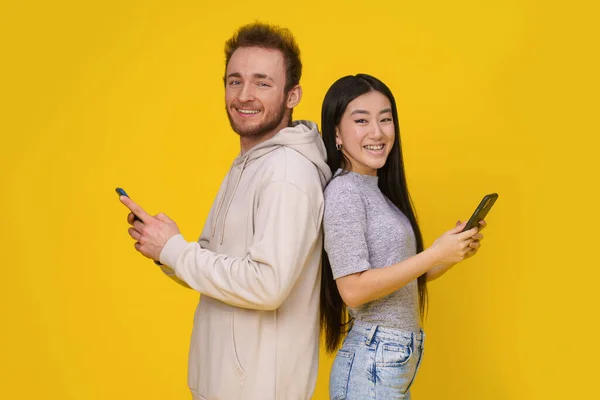 白种人和亚洲女孩背靠背站着 手里拿着智能手机 看着被黄色背景隔离的相机移动应用程序广告 产品摆放 — 图库照片