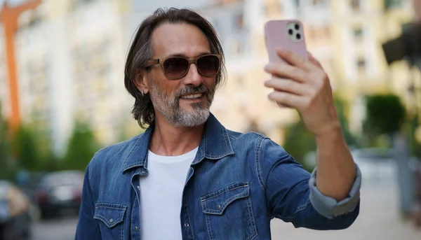 Handsome stylish sexy middle aged grey bearded man smiling on camera old town wearing casual. Happy senior man on street of european city making selfie using mobile phone. Travel concept.