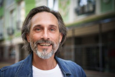 Happy handsome middle aged man standing outdoors on street of city looking at camera with a charming smile. Middle aged grey bearded man traveling in city smiling on camera wearing casual.  clipart