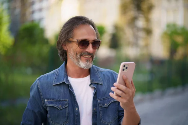 Happy adult man on street of european city making selfie using mobile phone. Travel concept. Handsome stylish sexy middle aged grey bearded man smiling on camera old town wearing casual.