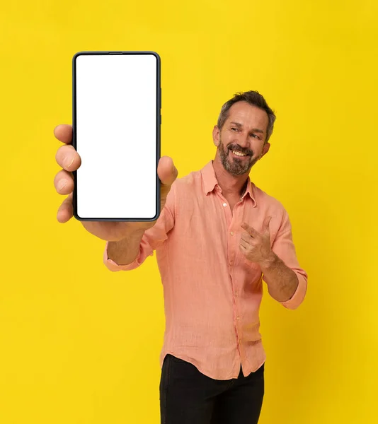 Happy middle aged grey haired fit man holding big smartphone pointing finger at it wearing peach shirt isolated on yellow. Mature muscled man with phone app advertisement. Mock up white screen.