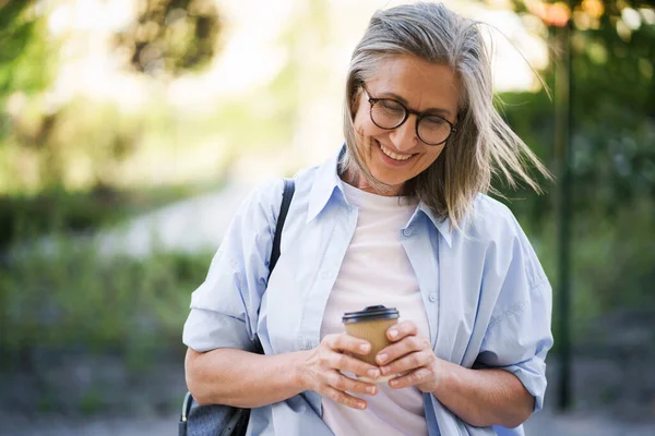 Mature woman tenderness. Charming mature woman drinks coffee on the go fixing her hair. Mature woman traveling enjoying cup of tea, coffee on the go. Grey haired woman in casual with backpack.