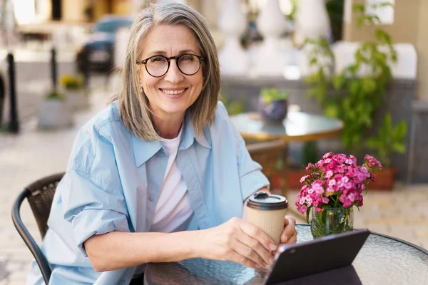 Mature European grey hair charming woman drink coffee using digital tablet sitting at table of street cafe. Mature silver hair woman working outdoors city cafe, wearing white t-shirt and blue shirt.