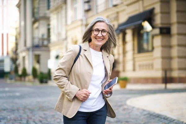 Mature elegant beautiful businesswoman. Senior woman in casual formal wear hold phone in hand walking on street from work or back to office. Mature woman spend free time traveling in European city.