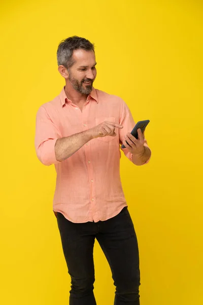 Middle aged grey haired handsome fit man holding smartphone looking at it wearing peach shirt isolated on yellow. Mature muscled man with phone, app advertisement.
