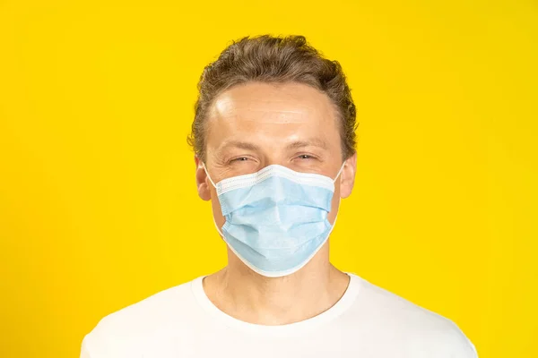 Happy, excited young man wearing a medical mask on his face. Sick young man in face mask wearing white t-shirt isolated on yellow background. A smiling caucasian man in protective face mask.