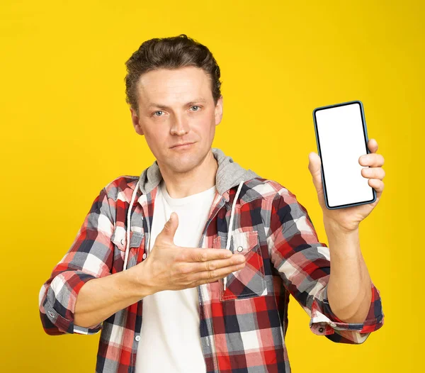 New app or website introduction holding smartphone with white screen blond man, wearing red plaid shirt. Man with phone display mock up isolated on yellow background. Mobile app advertisement.
