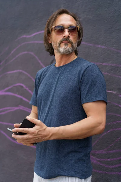 Confident mature man standing in front dark urban wall looking away with smartphone in hands wearing sunglasses, dark blue t-shirt and light shorts. Mature business man or freelancer outdoor.