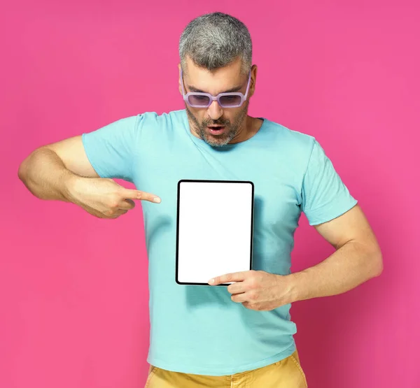 Pointing at digital tablet handsome man looking at white screen lowered his head wearing casual blue shirt and sunglasses isolated on pink background. Mobile app advertisement.